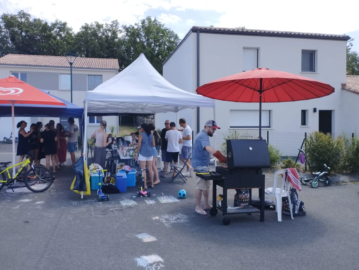 Voisins rassemblés dans la rue du lotissement pour la fête des voisins, sous les barnums et autour d'un barbecue.