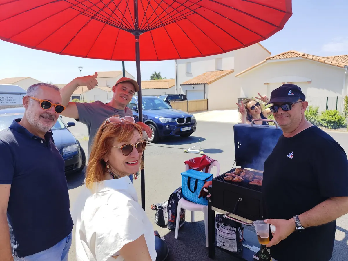 Groupe de voisins souriants autour du barbecue, sous un grand parasol rouge.